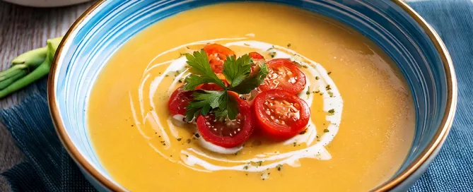 Blaue Schale mit oranger Suppe, garniert mit Kirschtomaten, Petersilie und einem Sahnewirbel, auf blauem Tuch mit Chicorée und Tomaten im Hintergrund.