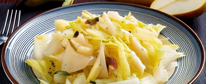 Teller mit Chicoreesalat, Birnenscheiben und Käse, im Hintergrund zwei ganze und eine halbierte Birne auf dunklem Untergrund.