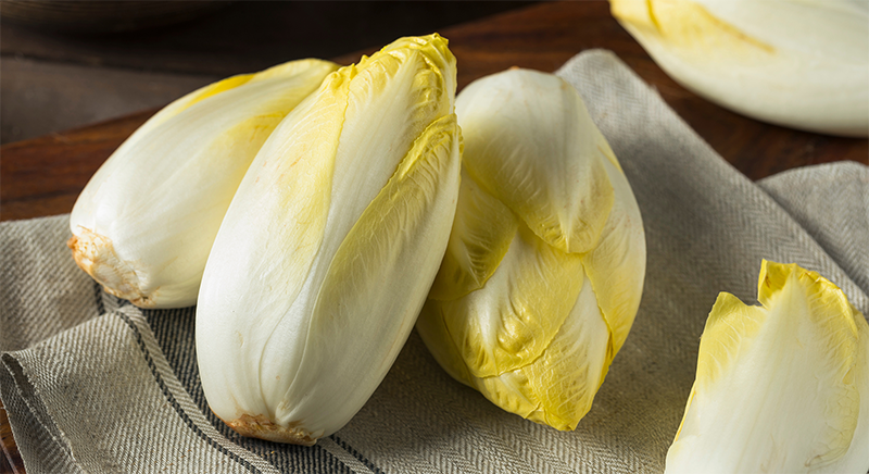 Drei frische, gelb-weiße Chicorée liegen auf einem Leinentuch auf einem Holztisch.