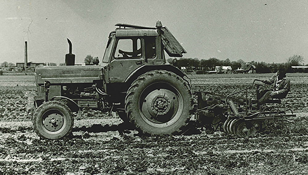 Alter Traktor mit großem Hinterrad und Pflug auf einem gepflügten Feld, im Hintergrund Bäume und Gebäude.