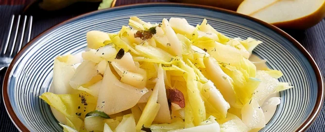 Teller mit Chicoreesalat, Birnenscheiben und Käse, im Hintergrund zwei ganze und eine halbierte Birne auf dunklem Untergrund.