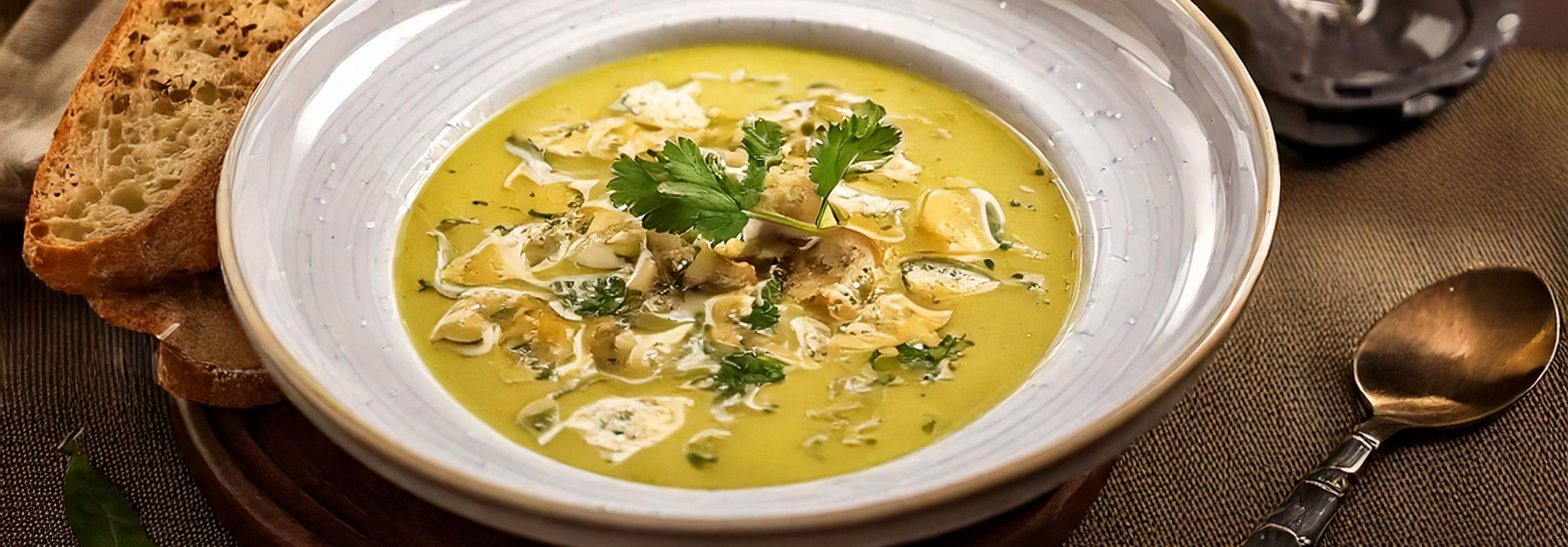 Schale mit gelber Chicorée-Suppe, garniert mit Petersilie und Mandelscheiben, umgeben von Knoblauch, Orange, Brot, Käse und Kräutern auf dunklem Untergrund.