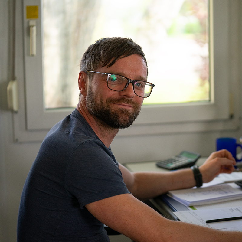 Mann mit Brille und dunkelblauem T-Shirt sitzt an einem Schreibtisch mit Unterlagen und einem Taschenrechner vor einem Fenster.