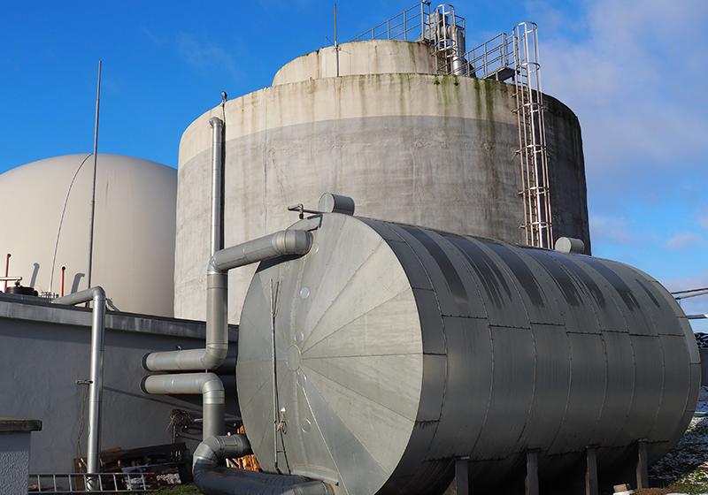 biogasanlage Große zylindrische und kugelförmige Behälter einer Biogasanlage mit Leitern und Rohren unter blauem Himmel.