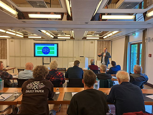 chicoree-tagung_2025_vorträge_sl_gartenbau_web Mehrere Personen sitzen an Tischen in einem Tagungsraum und hören einem stehenden Vortragenden zu, der vor einem Bildschirm mit Diagramm spricht.