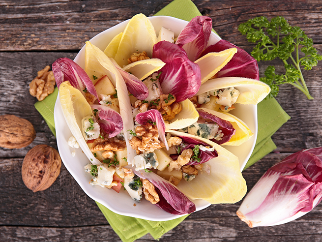 roter_chicorée_wissenswertes_salatidee_web Weißer und roter Chicorée in einer Schale mit Walnüssen und Kräutern auf grünem Tuch und Holztisch.