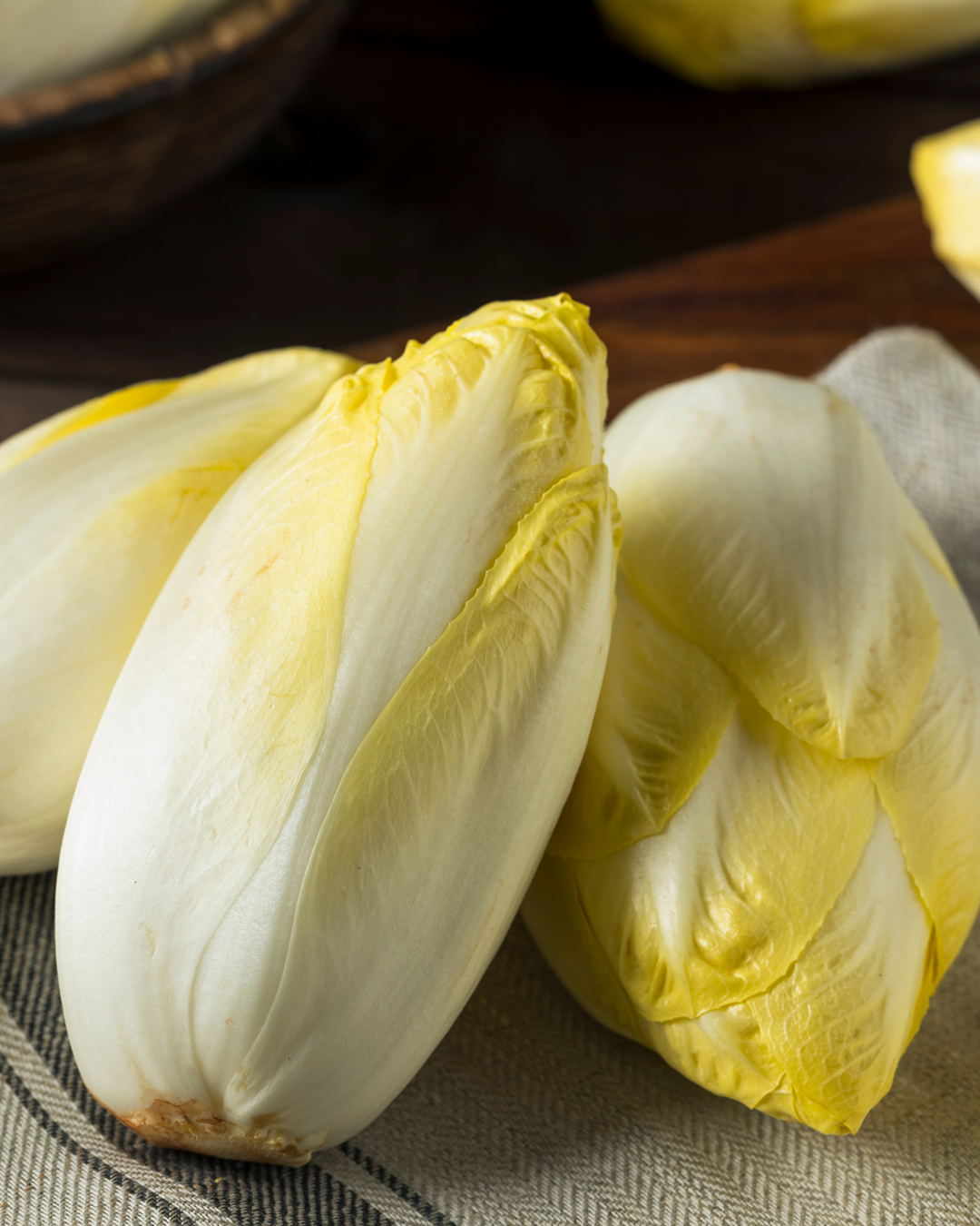 gesundes_aus_schwanteland_header_mobil Drei frische, gelb-weiße Chicorée liegen auf einem Leinentuch auf einem Holztisch.