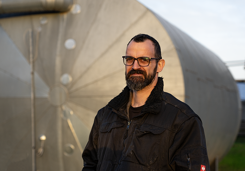 Johann_Stueben Mann mit schwarzer Jacke steht vor einer Biogasanlage mit großen silbernen Behältern im Hintergrund